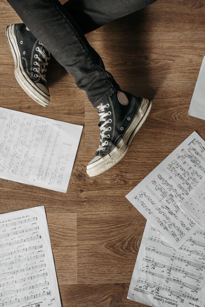 Top view of person in black sneakers with sheet music scattered on wooden floor.